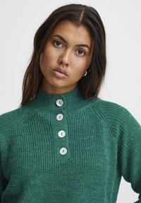 Groene gebreide trui met hoge kraag, vijf knoopsluitingen aan de voorkant en een textuurpatroon, met raglanmouwen.