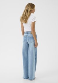 Woman with long blonde hair wearing a white short-sleeve top and light blue wide-leg jeans, standing against a plain white background.