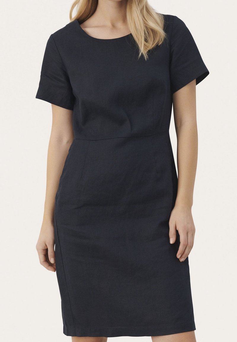 Femme portant une robe noire ajustée à manches courtes, longueur genou, avec un col arrondi, debout devant un fond clair uni.