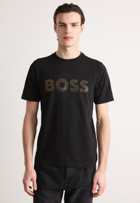 Juodas medvilnės T-shirt su apvaliu kaklu, ant krūtinės spausdinamas "BOSS" aukso spalva. Trumpi rankoviai, reguliariai prigludimas.