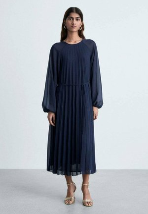 Navy blue pleated dress with long sleeves, round neckline, and a tie waist detail. Sheer fabric creates a light texture. Gold sandals.