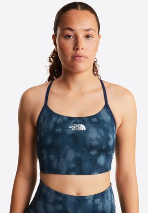 Kobieta z kręconymi włosami związanymi do tyłu, ubrana w granatowy sportowy biustonosz w kwiaty oraz pasujące wysokiej talii legginsy z logo The North Face.