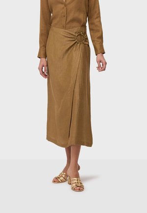 Kobieta w brązowej, długorękawowej sukience zapinanej na guziki, z marszczonym detalem na wysokości talii, w połączeniu ze złotymi sandałami na obcasie z odkrytymi palcami.
