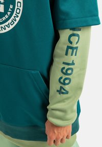 Bluza z kapturem w kolorze niebieskozielonym i jasnej zieleni. Posiada duży graficzny logo oraz napis "ICE 1994" na rękawie. Miękka tekstura, swobodny fason.