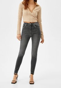 Jeans skinny altos de cintura en gris con detalles desgastados claros y costuras en contraste, combinados con un suéter corto de espiga en color beige.