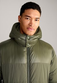 Gepolsterte olivgrüne Jacke mit hohem Kragen, Reißverschluss vorne, Druckknöpfen und kontrastierenden Texturpanelen. Glatte, glänzende Stoff.
