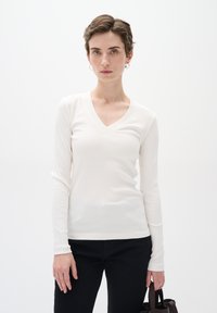Weißes Langarm-V-Ausschnitt-Oberteil mit gerippter Textur, kombiniert mit schwarzen Hosen. Das Model hält eine kleine braune Handtasche und steht vor einem schlichten Hintergrund.
