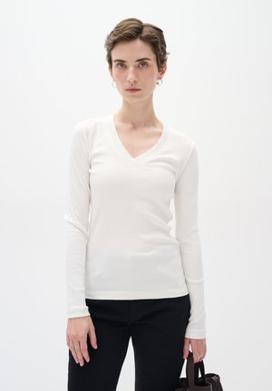 Weißes Langarm-V-Ausschnitt-Oberteil mit gerippter Textur, kombiniert mit schwarzen Hosen. Das Model hält eine kleine braune Handtasche und steht vor einem schlichten Hintergrund.