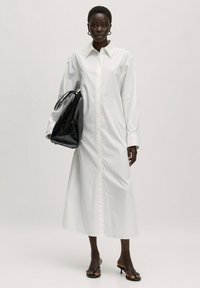 Weißes langärmliges Hemdkleid mit Knopfleiste und spitzem Kragen, kombiniert mit einer schwarzen glänzenden Handtasche und schwarzen Sandalen mit Riemchen.