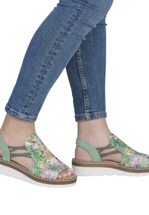 Personne portant des sandales ouvertes vertes à motifs floraux avec des brides élastiques et des semelles blanches, associées à un jean skinny bleu retroussé.