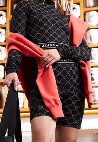 Svart långärmad klänning med smiley-ansiktsmönster och figursydd design, ihop med en korallfärgad sweatshirt som hänger över axlarna.