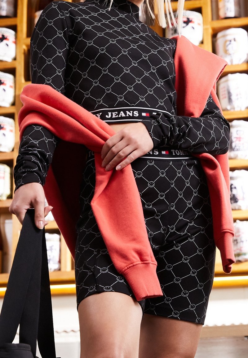 Svart långärmad klänning med smiley-ansiktsmönster och figursydd design, ihop med en korallfärgad sweatshirt som hänger över axlarna.