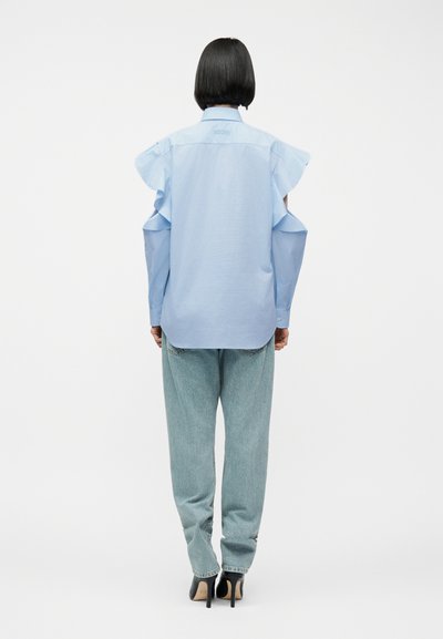 Personne aux cheveux noirs courts portant un chemisier bleu clair à épaules dénudées, un jean bleu clair et des chaussures à talons noires, debout de dos.