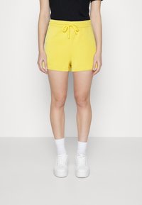 Shorts en coton jaune avec une ceinture élastique et un cordon de serrage. Associés à des baskets blanches et des chaussettes blanches jusqu'à la cheville.