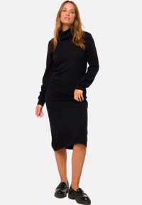 Robe noire à manches longues avec un col drapé et une silhouette ajustée, présentant un ourlet à hauteur de genou et une texture douce. Portée avec des chaussures noires.