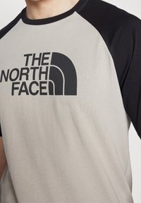 Camiseta de hombre con un cuerpo gris y mangas negras, cuello redondo y un destacado diseño de logo negro en el frente.