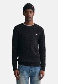 Schwarzer Kabelstrickpullover mit Rundhalsausschnitt und gerippten Bündchen, kombiniert mit blauer Jeans. Besitzt ein kleines Logodetail auf der Brust.