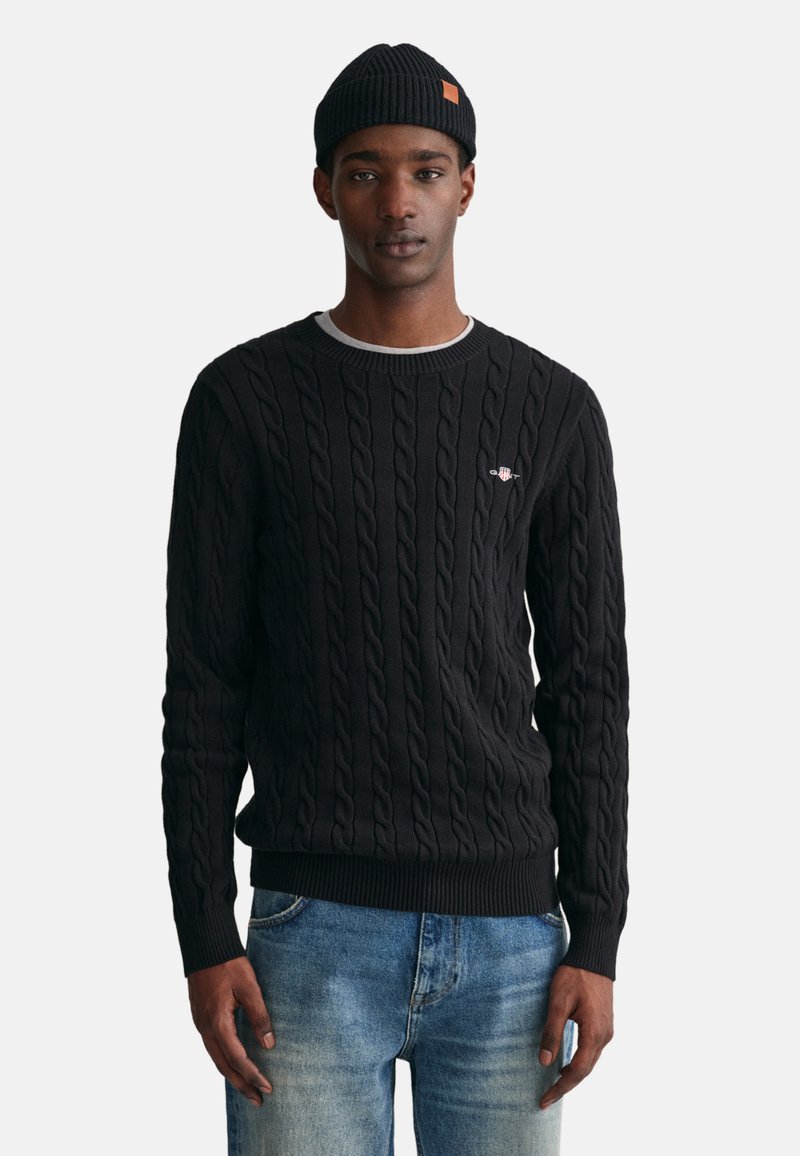 Schwarzer Kabelstrickpullover mit Rundhalsausschnitt und gerippten Bündchen, kombiniert mit blauer Jeans. Besitzt ein kleines Logodetail auf der Brust.