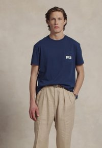 T-shirt de algodão azul-marinho com um bolso exibindo o logótipo "POLO", combinado com calças claras de linho plissadas, apresentando um cinto e um relógio.