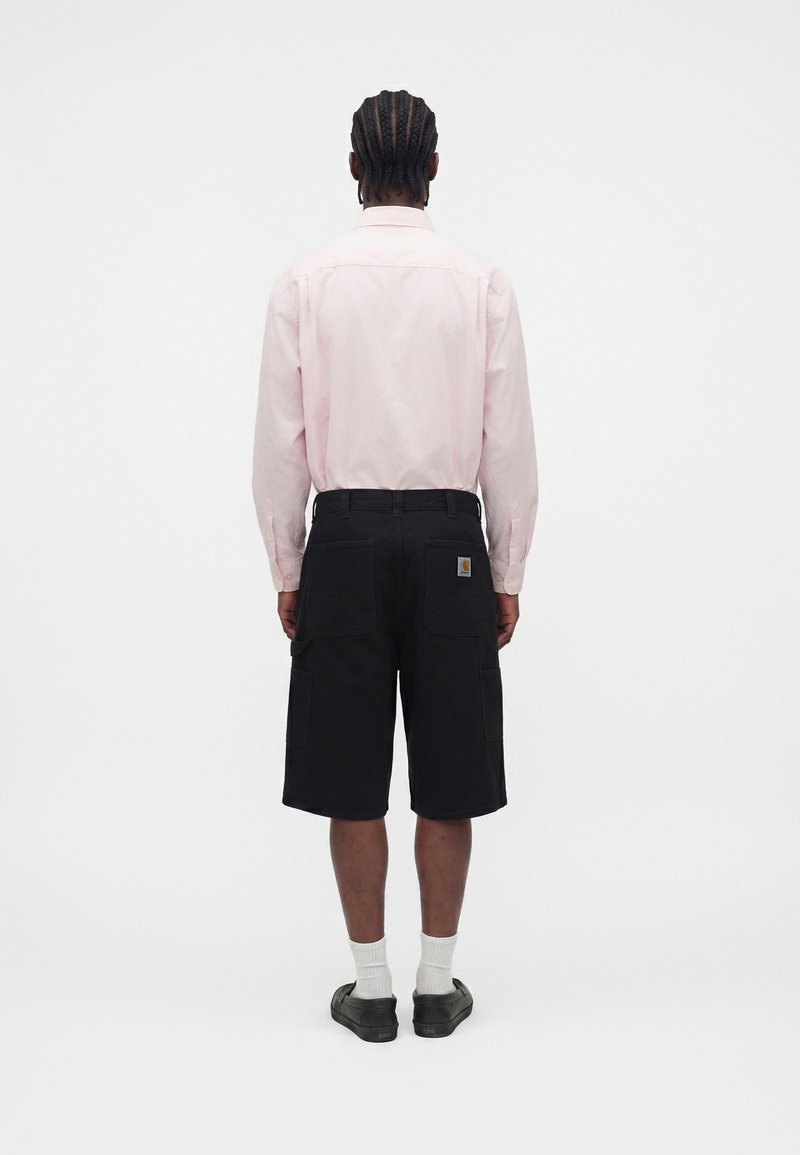 Mann mit geflochtenem Haar, der ein hellrosa langärmliges Hemd, schwarze kniehohe Cargoshorts, weiße Socken und schwarze Schuhe trägt, steht mit dem Rücken zur Kamera.