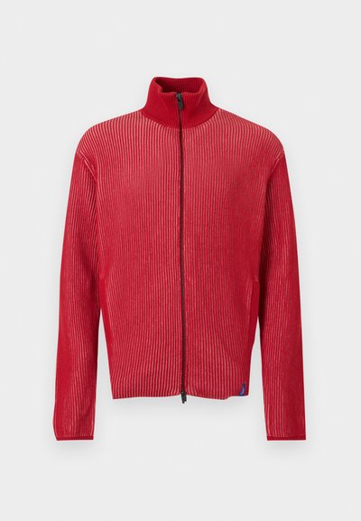 Κόκκινο ριμπ πλεκτό πουλόβερ με φερμουάρ, ψηλό γιακά και μακριά μανίκια, εμφανίζεται μπροστά σε απλό φωτεινό φόντο.