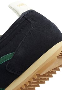 Schwarzer Wildlederschuh mit grünen Akzenten, einer beigen Gummisohle und genähten Details im Obermaterial. Verfügt über eine weiche, gepolsterte Ferse.