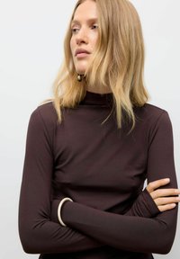 Maglia a maniche lunghe e collo alto in tessuto marrone scuro; silhouette aderente con dettagli per foro per il pollice; indossata con un bracciale al polso.