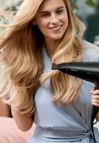 Ein schwarzer Haartrockner, der in der rechten Hand gehalten wird, ein Model mit langen, welligen blonden Haaren, das einen hellblauen Bademantel trägt und lächelt, in einem Innenraum mit Pflanzen.