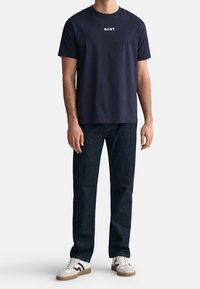 T-shirt di cotone blu navy con collo a giro e logo bianco "GANT" sul petto, abbinata a jeans in denim scuro e sneakers bianche con dettagli neri.