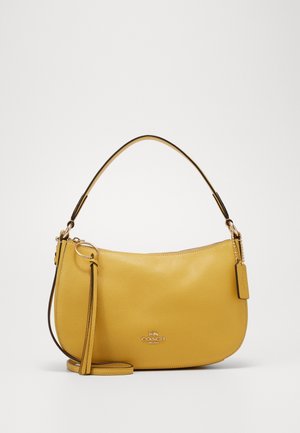 Gele leren Coach schoudertas met gouden hardware, ritssluiting, kwastdetail en een afneembare riem op een witte achtergrond.