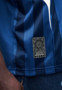 Κοντινό πλάνο μπλε ριγωτού υφάσματος με μια μαύρη ετικέτα «Authentic Lifestyle Motorsport», φορεμένο πάνω από σκούρο τζιν παντελόνι.