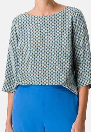 Kobieta w luźnej, wzorzystej bluzce z niebieskimi geometrycznymi kształtami i jasnych niebieskich spodniach z wysokim stanem.