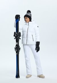 Doudoune blanche avec accents noirs, pantalon de ski blanc et gants noirs. Tiens un ski bleu avec des fixations noires, portant un bonnet noir à motifs.