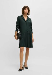 Frau trägt ein dunkelgrünes langärmliges Kleid, schwarze High Heels, eine goldene Halskette und hält eine schwarze Handtasche vor einem einfarbigen Hintergrund.