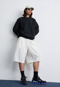 En person står op ad en hvid væg iført en sort sweatshirt, hvide shorts, solbriller og en beige kasket, hvilket fremviser en afslappet stil.