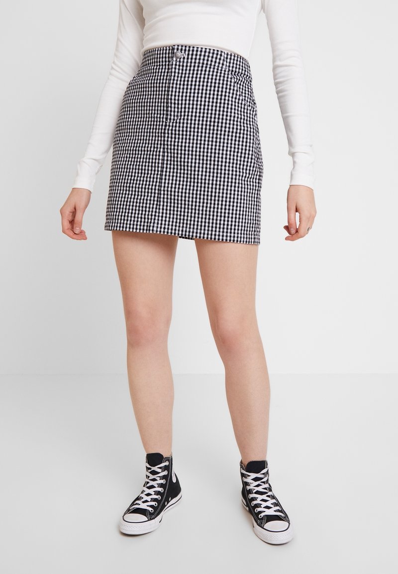 Jupe mini en vichy noir et blanc avec une taille cintrée, une fermeture à boutons sur le devant et un devant plat. Portée avec des baskets montantes noires.