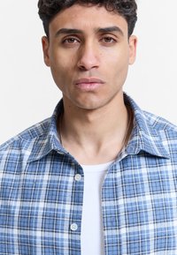Jeune homme aux cheveux courts et bouclés portant une chemise à carreaux bleue par-dessus un haut blanc, regardant droit devant avec une expression neutre.