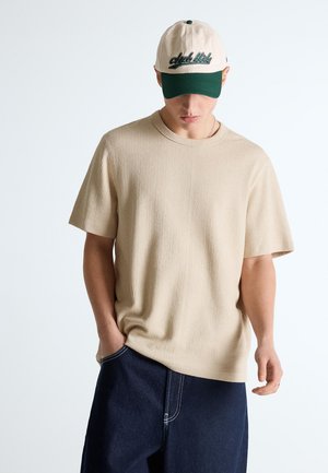 Férfi bézs texturált rövid ujjú ingben, sötétkék nadrágban és bézs-zöld baseball sapkában, amin "club ltd." felirat olvasható, lefele néz, kezét zsebében tartja.