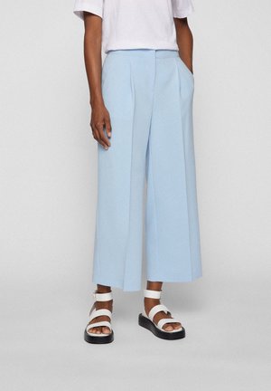 Person, die hellblaue, weit geschnittene, verkürzte Hose, weißes T-Shirt und weiße Sandalen mit schwarzen Sohlen trägt, stehend vor einem schlichten Hintergrund.