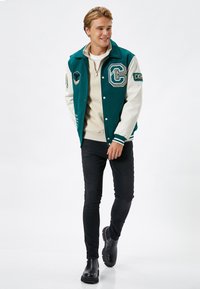 Teal-tyylinen yliopistojakku, jossa on valkoiset hihnat ja brodeeratut patchit, yhdistettynä kermaiseen neuleeseen ja mustiin kapeisiin farkkuihin, viimeistelty mustilla saappailla.