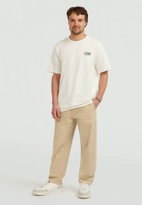 Homem vestido com uma t-shirt O'Neill off-white, calças bejes e ténis brancos, em pé com uma mão no bolso contra um fundo claro e liso.