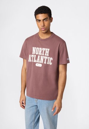 Joven con camiseta color malva estampada con "North Atlantic" y jeans azul claro, posando frente a un fondo claro y liso.