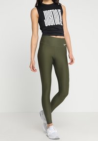 Top sin mangas negro con el texto "JUST DO IT", combinado con leggings verde oliva de cintura alta. El calzado son zapatillas blancas. Diseño simple y deportivo.