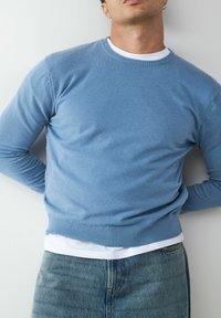 Jasnoniebieski sweter z dzianiny z okrągłym dekoltem, prążkowanymi mankietami i dołem, noszony na białym t-shircie, w zestawie z dżinsami.