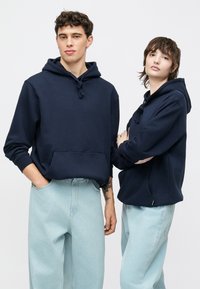 Tamsiai mėlyni džemperiai su gobtuvu, priekyje kišene, su raišteliais ir ribotom rankogaliais; derinami su šviesiai mėlynomis laisvo kirpimo džinsais. Paprastas dizainas; minkšta tekstūra.