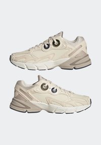 Beige sportskor med texturerad syntetisk ovandel, vadderad krage, runda snörhål och kontrasterande grå detaljer på sålen och hälen.