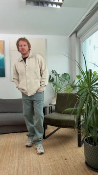 Beige genser-skjortejakke, grønne jeans og grå joggesko. Innendørs med planter, en mørk sofa og en stol med grønn pute.