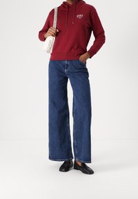 Sudadera con capucha burdeos con logotipo frontal, combinada con jeans de mezclilla azul de pierna ancha. Usada con zapatos negros tipo slip-on y un bolso beige.