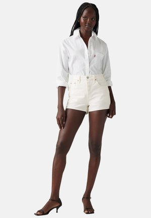 Camicia bianca con bottoni, shorts di denim bianchi a vita alta e sandali neri con tacco. Caratteristiche includono una tasca frontale e accessori in bronzo.