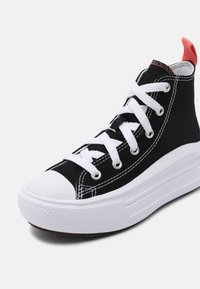 Baskets montantes noires avec semelle blanche épaisse, lacets blancs, bout de la chaussure blanc et languette rouge à l'arrière, orientées vers la droite.